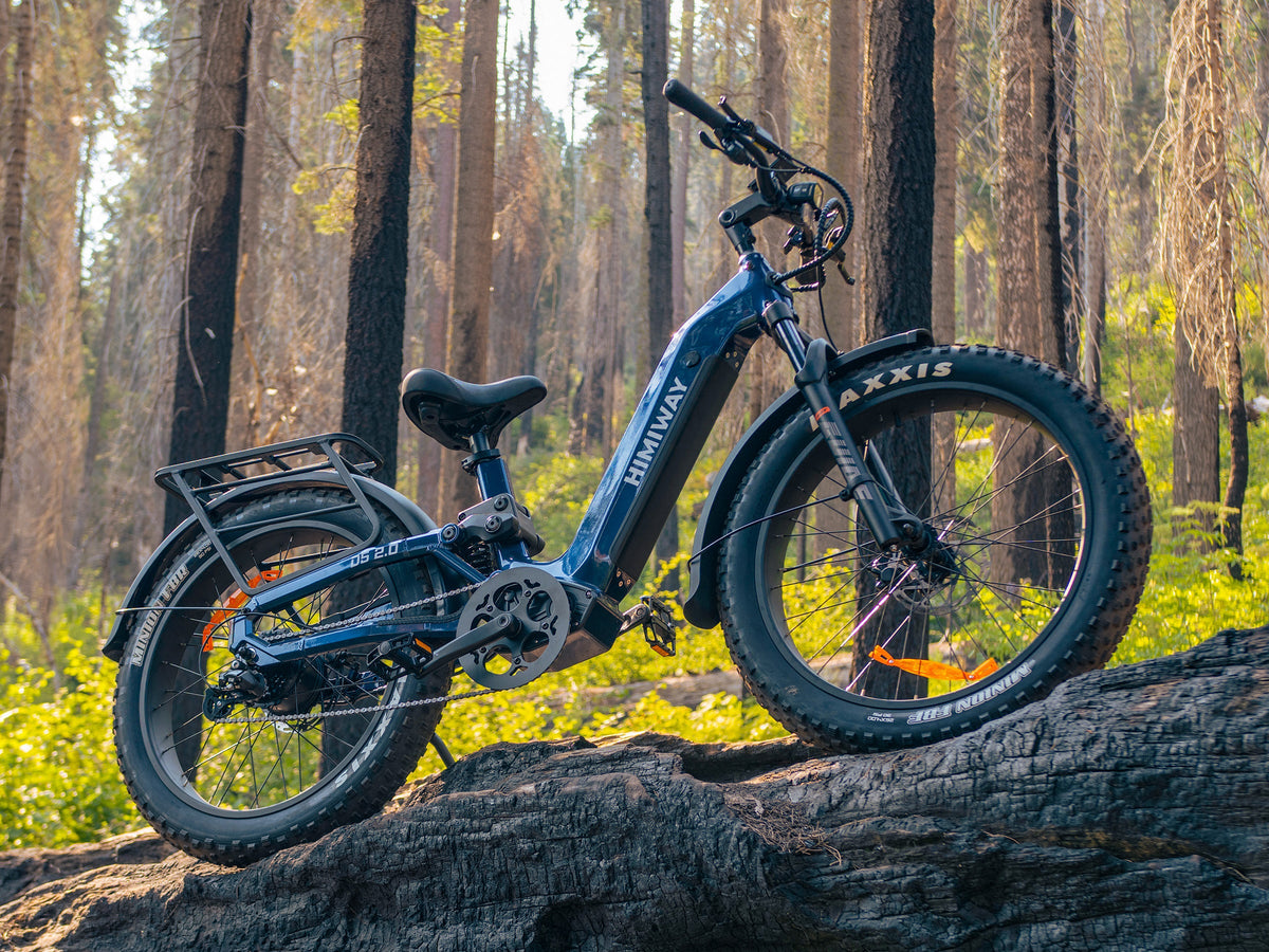 A Midnight Blue Himiway D5 2.0 step-thru fat tire electric bike parked on a large fallen log in a dense forest.